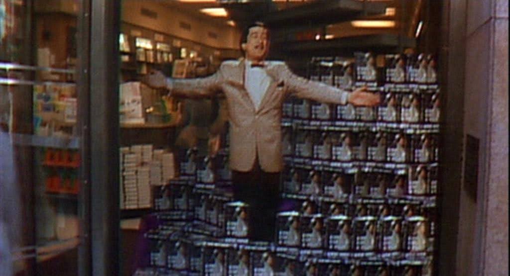 A cardboard cutout of Rupert Pupkin (Robert DeNiro) in a tan suit, black pants, red bow tie, arms outstretched, in a shop window advertising a stack of his book, The King of Comedy (1982).