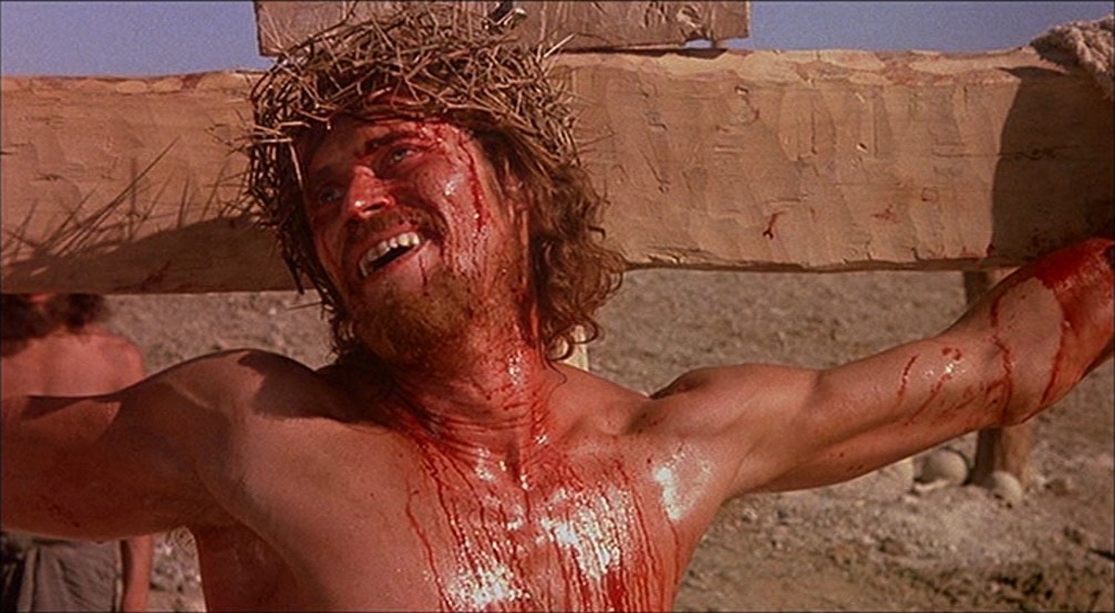 Jesus (Willem Dafoe), shirtless, dangling from the cross, covered in blood, wearing a crown of thorns and smiling in the moments before death, from The Last Temptation of Christ (1988).