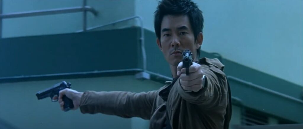 A man in a brown jacket points two guns, one off to the right and one at the screen. Behind him is a building with a dark roof.