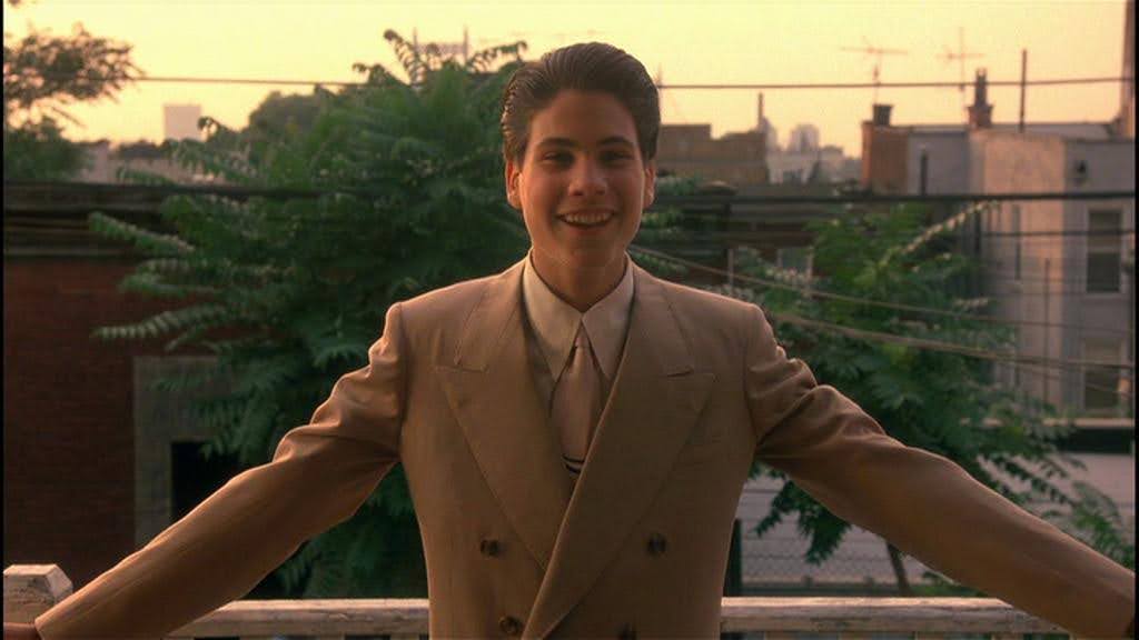 Young Henry Hill (Christopher Serrone) trying on a new tan suit, smiling towards people off camera, backlit by sunset over the Brooklyn skyline, from Goodfellas (1990).