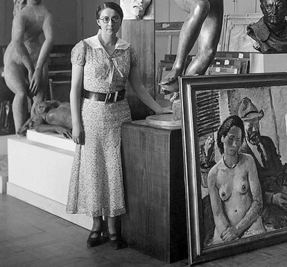 black-and-white image of a white woman with dark hair standing amid several sculptures and one painting