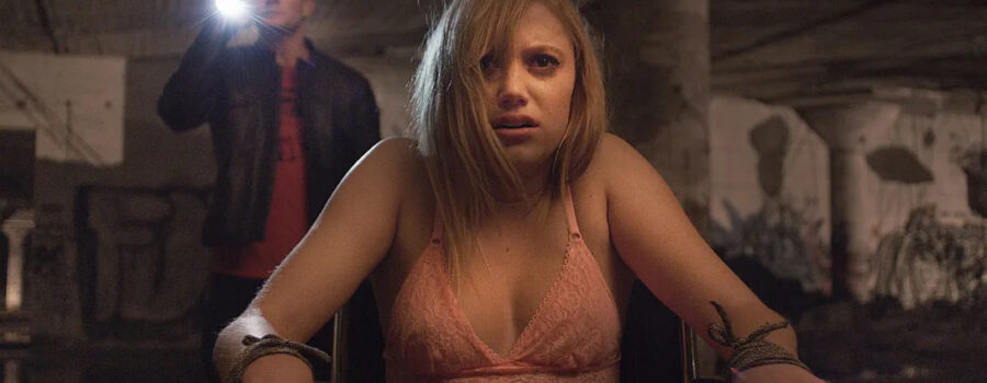 Jay (Maika Monroe) is tied to a wheelchair in her bra and underwear looking on in horror at It off camera as Jeff (Jake Weary) stands behind her with a flashlight on It so she can see what he is explaining.