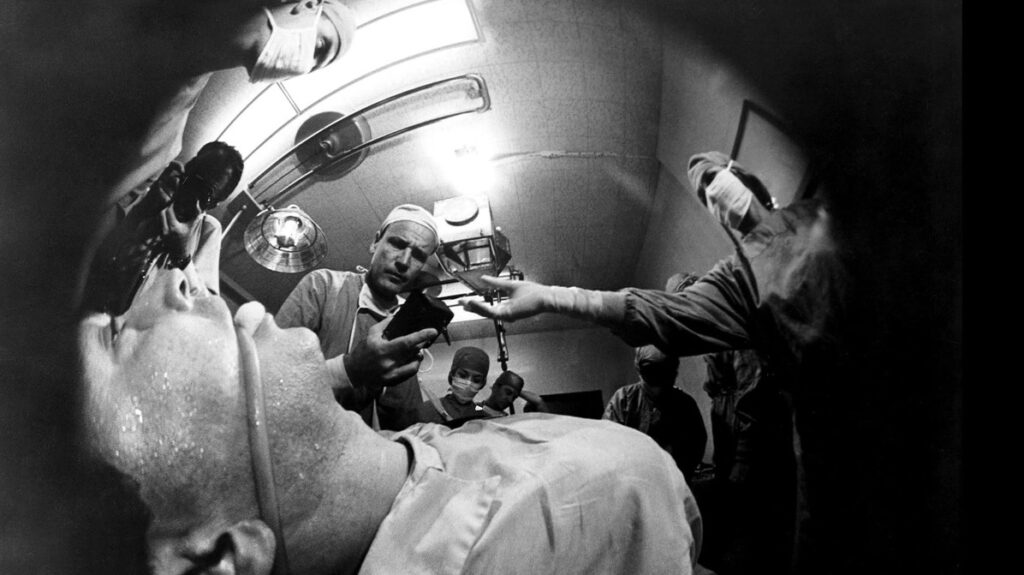 A low fish-eye angle of a man strapped to a gurney. He is surrounded by individuals in surgical garments, There is a tube wrapped around his mouth.