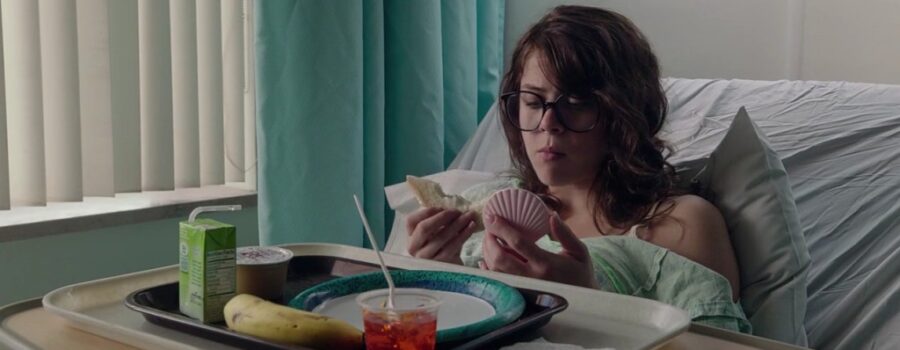 Olivia Luccardi as Yara Davis, a young long-haired white brunette wearing large black-rimmed glasses and a teal hospital gown, sits up on a hospital bed at a tray with a plate, juice box, jello cup, applesauce, and a banana. In one hand is a sandwich on white bread with a bite taken out of it, and in the other is her pink clamshell e-reader, which is she is staring into. A wrinkled periwinkle pillow and bed sheet are behind her, as are white vertical blinds and a calm blue curtain.