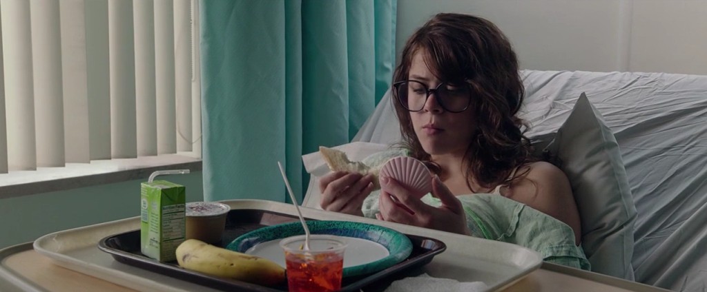 Olivia Luccardi as Yara Davis, a young long-haired white brunette wearing large black-rimmed glasses and a teal hospital gown, sits up on a hospital bed at a tray with a plate, juice box, jello cup, applesauce, and a banana. In one hand is a sandwich on white bread with a bite taken out of it, and in the other is her pink clamshell e-reader, which is she is staring into. A wrinkled periwinkle pillow and bed sheet are behind her, as are white vertical blinds and a calm blue curtain.