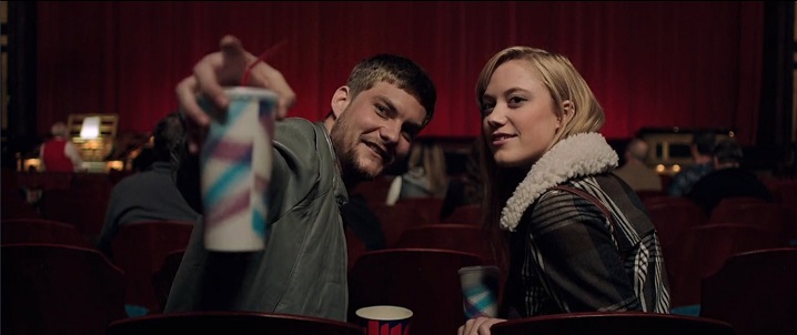 Jake Weary as Jeff, a scruffy young white man with brown hair and wearing an olive jacket on the left, and Maika Monroe as Jay, a young white blond woman with pink lipstick and wearing a leather bomber jacket on the right, are in vintage movie theater seats, turned around and looking into the distance. Jeff is pointing to the upper left with a generic fountain soda cup in his hand. Both have happy, interested looks on their faces. In the background is the red velvet curtain in front of the screen and an out-of-focus organist to the left.