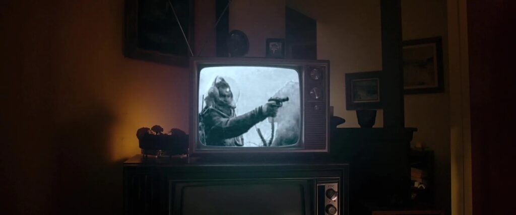 A late-70s, early-80s style square television sits in center frame atop an older, larger early-70s style television entertainment center. The smaller, newer TV shows a black-and-white still of a retro-futurist astronaut holding a gun-like weapon while wearing a spacesuit adorned with a comically oversized clear helmet. In the background of the shot is a low-level yellow and assorted silhouetted decor: picture frames, flowers, a ledge and room divider.