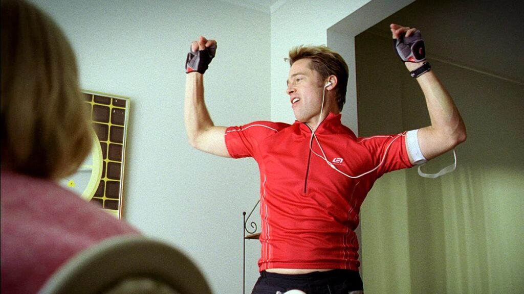 A man wearing a tight fitting cycling outfit (including a red shirt, gloves, and MP3 player armband) dances with his hands above his head. 