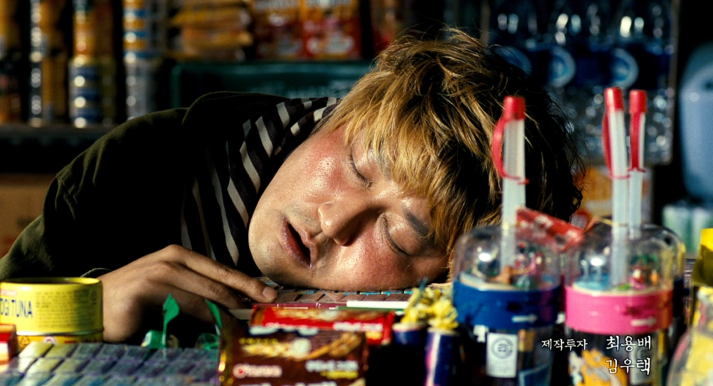 A man lays asleep at the window to a food cart. His cheek is resting on a pile of quarters and candy.