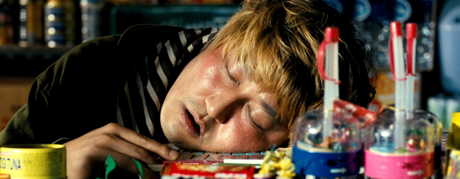 A man lays asleep at the window to a food cart. His cheek is resting on a pile of quarters and candy.