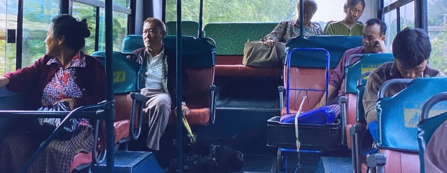 The Outsider, an old Japanese man, sits apart from others on a city bus with four black chickens tied together near his feet.