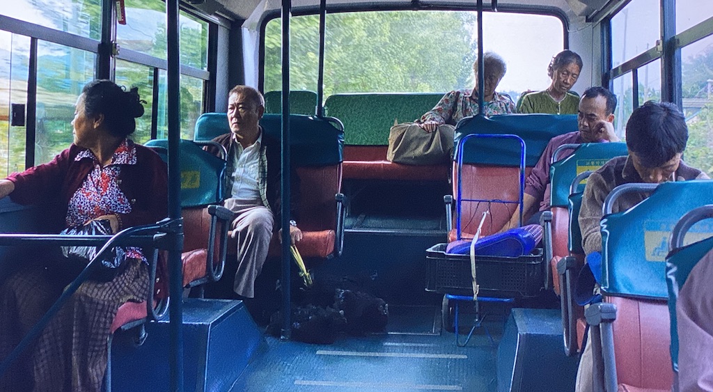 The Outsider, an old Japanese man, sits apart from others on a city bus with four black chickens tied together near his feet. 