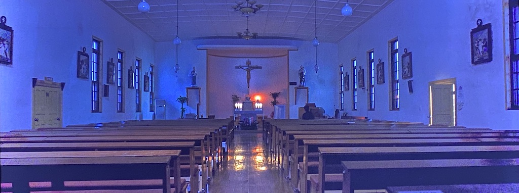 Image of a Catholic church at night with several empty pews, an altar and a large crucifix. A lone priest sits in the front row. 