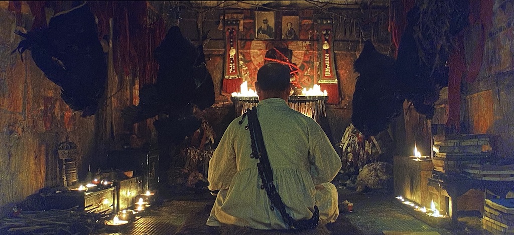 The outsider sits before his altar with his back to camera, surrounded by candles and black chickens hanging from the ceiling.