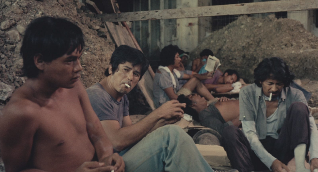 Several construction workers sitting and taking a break from work where a few workers are seen smoking a cigarette, one is seen reading a newspaper, and some are napping.