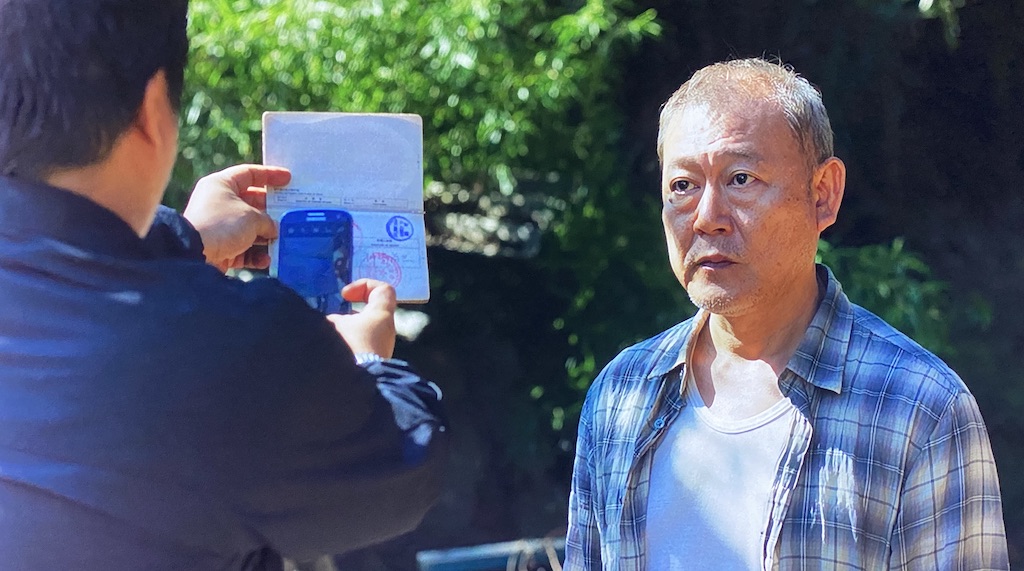 Officer Gong-ju takes a photo of The Outsider’s passport as the Outsider looks on. 