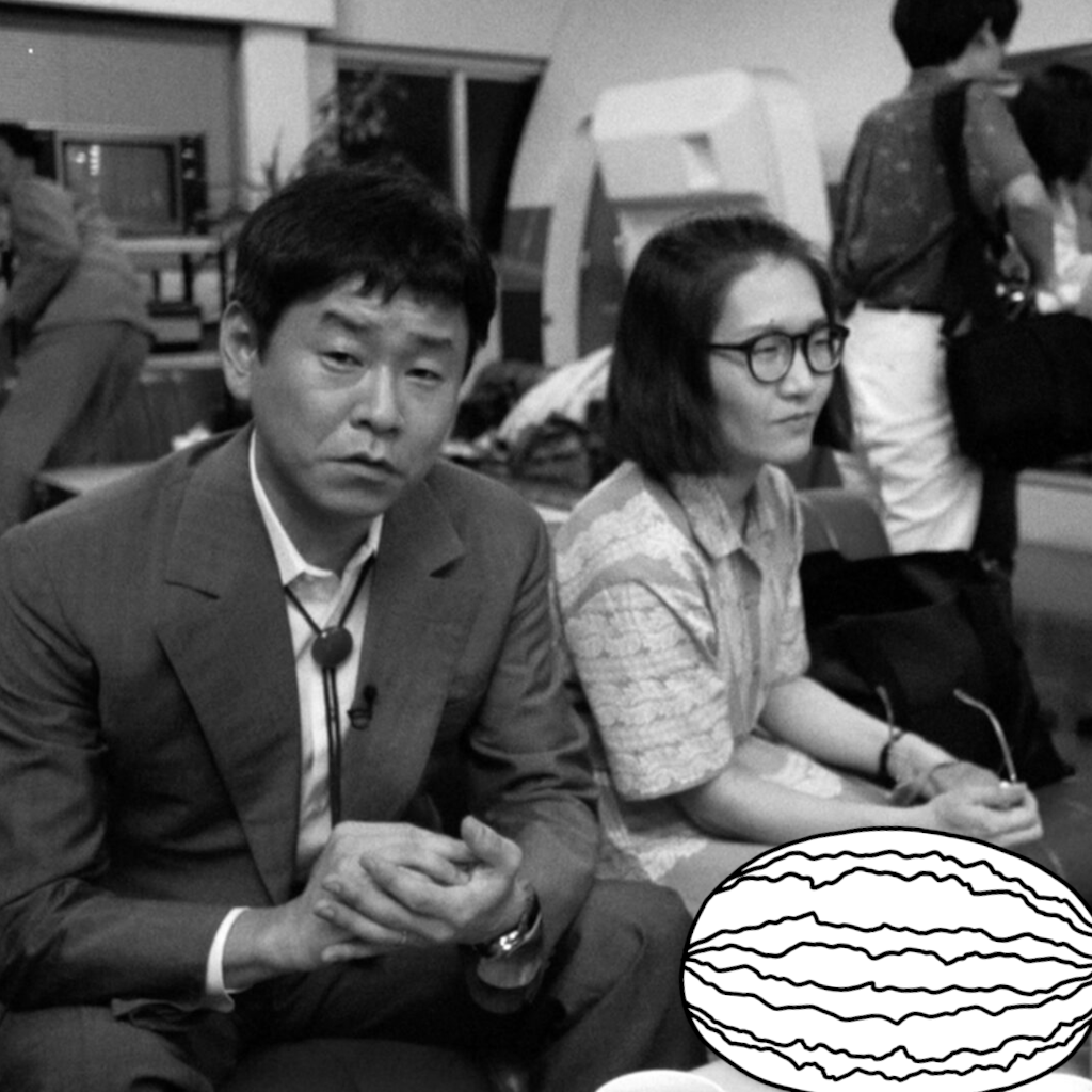 A man and a woman are sitting on a bench in an airport. The man is looking at the camera. On the table in from of them is a drawn watermelon.