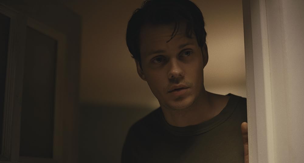 A light-skinned man with short brown hair and dark eyes stands in a white door frame. The background shows a ceiling illuminated with warm light from below.