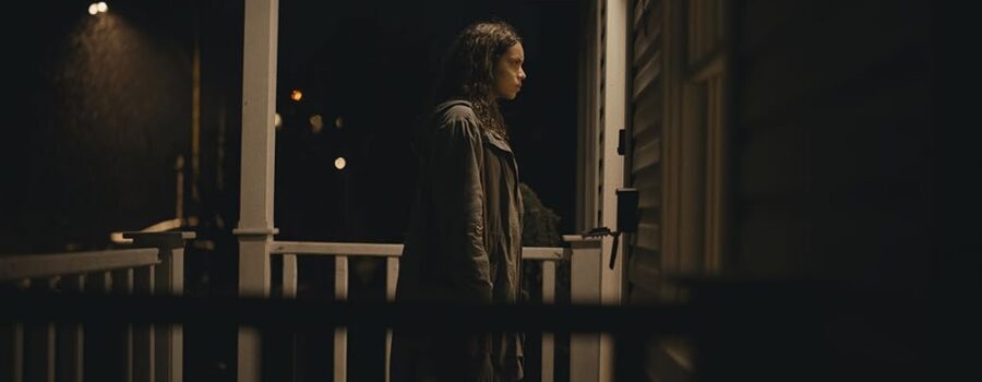 A brown-skinned woman with medium long, loose hair stands on an illuminated front porch of a house, facing the door. It's dark and raining outside, and the woman's hair and coat are wet.