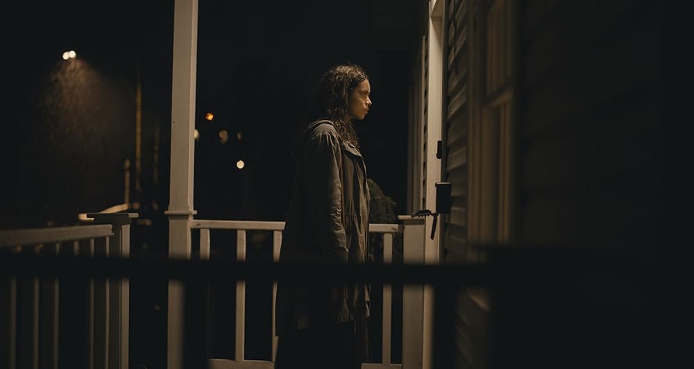 A brown-skinned woman with medium long, loose hair stands on an illuminated front porch of a house, facing the door. It's dark and raining outside, and the woman's hair and coat are wet.