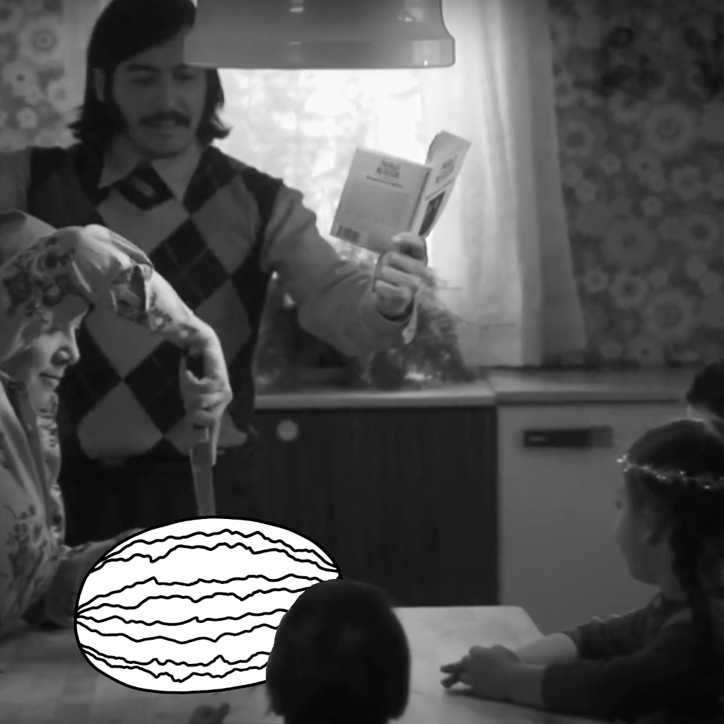 There is a table in the foreground of a kitchen. A man is standing behind the table holding a book. A woman is holding a knife and cutting a drawn watermelon. Three children are sitting around the table. Everyone is looking at the watermelon.