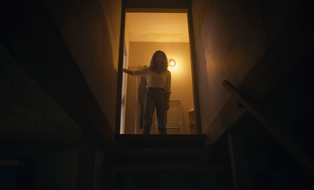 A photograph of a young woman mostly in silhouette looking down a dark stairwell. She is wearing a white blouse and blue jeans and has her right hand on the doorframe.