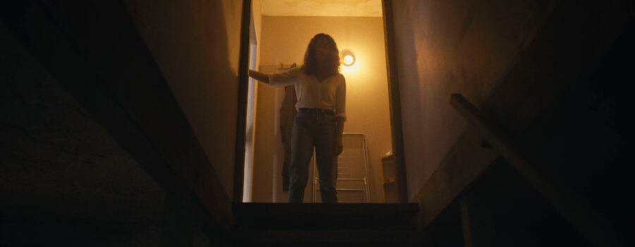 A photograph of a young woman mostly in silhouette looking down a dark stairwell. She is wearing a white blouse and blue jeans and has her right hand on the doorframe