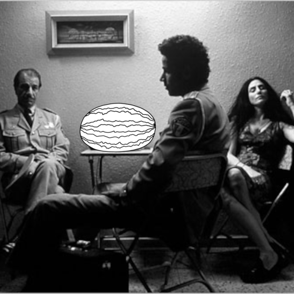 Two men and a woman are sitting around a table with a drawn watermelon on it. The older man is looking at the younger one. The woman's eyes are closed.