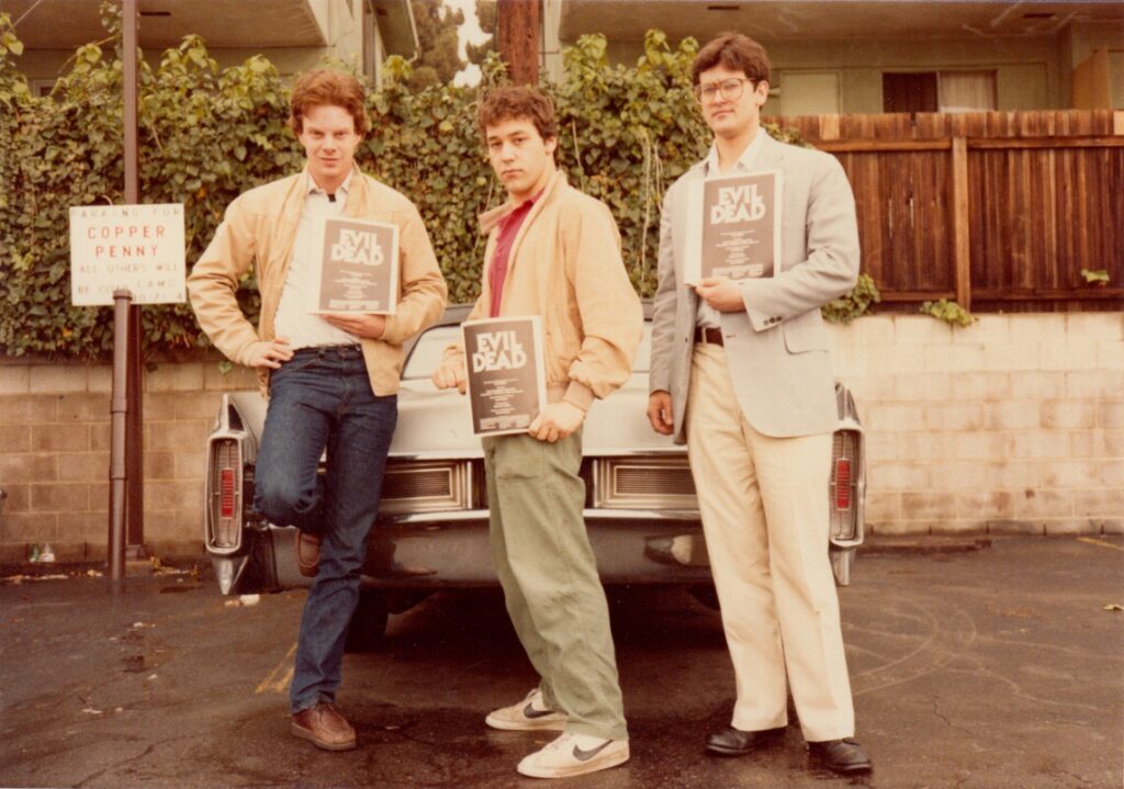 Three men stand in front of a car holding packets reading Evil Dead