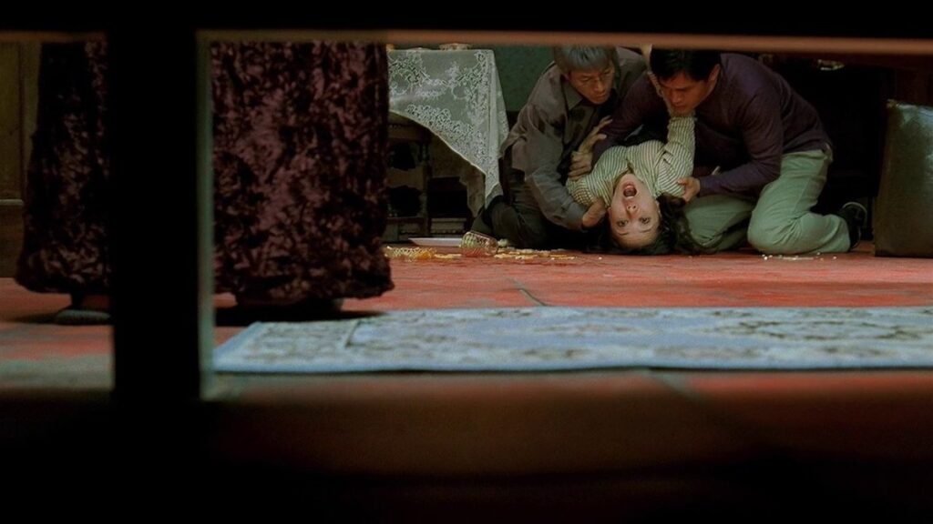 A low shot from the vantage point of under a piece of furniture, which frames the view, two men are seen cradling the flailing body of a woman, whose back is arched and looking out and above the lens. Dishes and pills are scattered on the burnt orange floor, where there is also a lace-covered table in the background, a velour-covered table in the middle ground, and a blue-gray Persian rug in the foreground.