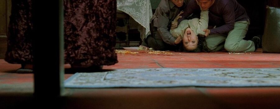 A low shot from the vantage point of under a piece of furniture, which frames the view, two men are seen cradling the flailing body of a woman, whose back is arched and looking out and above the lens. Dishes and pills are scattered on the burnt orange floor, where there is also a lace-covered table in the background, a velour-covered table in the middle ground, and a blue-gray Persian rug in the foreground.