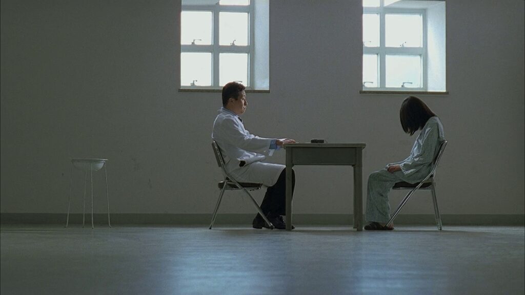 A side view of two figures in metal chairs slightly off-center in an all-white room with two medium-sized four-pane windows with handles. Between the two figures is a small waist-high table. The figure on the left resembles a male doctor in a white coat, hands on the table in front of him, his eyes aimed at the figure across from him, who resembles a young woman in a hospital gown, arms at her sides, facing down with long, black hair obscuring her face. There is a book on the table. Also in the room to the left is a white bowl set in a wire frame, elevated off the ground.