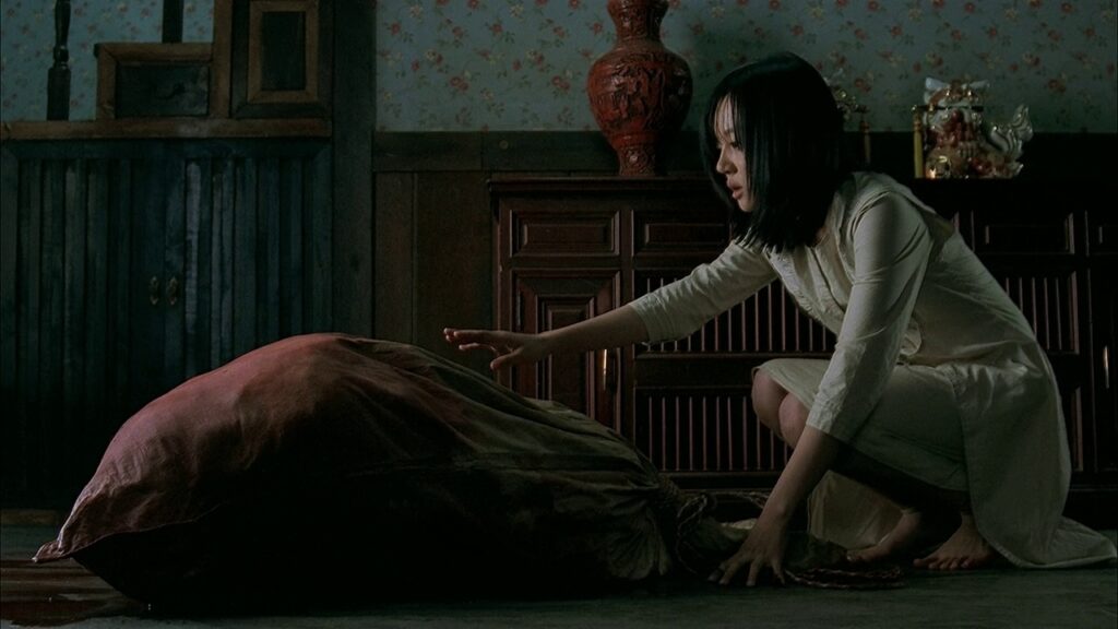 A two-thirds side view of a young barefoot woman on the right side of the shot and a large burgundy fabric sack on the left. The woman is crouched down with a hand outstretched toward the sack and her other hand holding her balance on the floor. She is wearing an off-white dress, has medium-length black hair, and an anxious look on her face. Between the woman and the sack are (from left to right) a partially out-of-view staircase, dark brown wainscotting with light floral wallpaper complementing it, and a large dark wooden dresser, intricately designed with a reddish vase and other ornamental items on top.