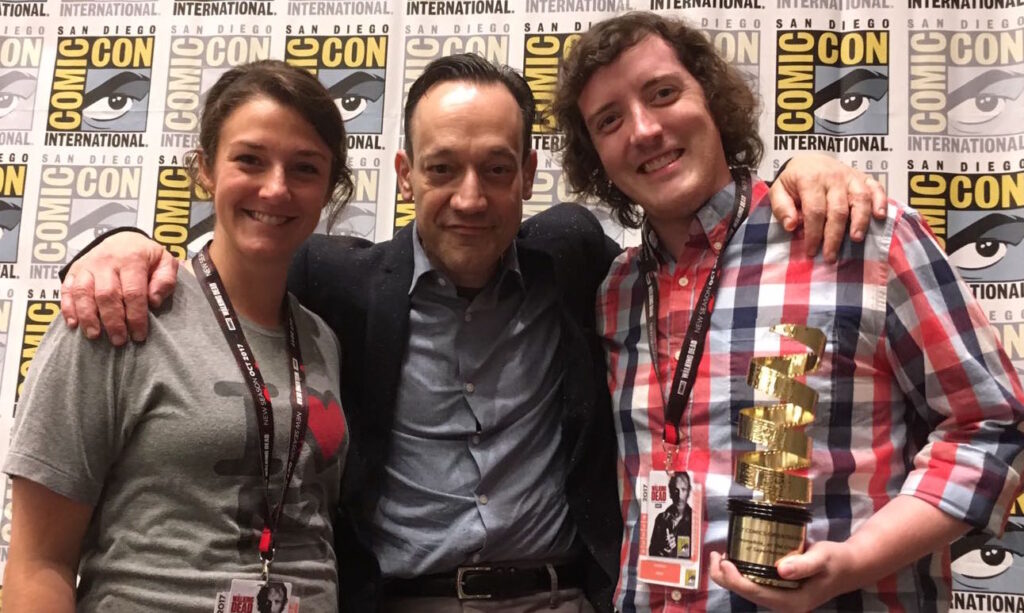 A woman in a gray shirt, a man in a sport coat, and a man in a plaid shirt stand in front of a convention backdrop.