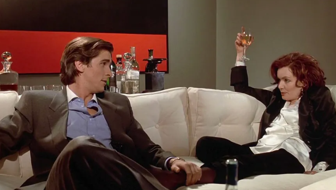 Guinevere Turner as Elizabeth sits on a couch next to Christian Bale as Patrick Bateman