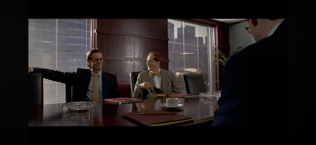 Patrick Bateman and Luis Carruthers sit at a table in a classy, wood paneled boardroom.