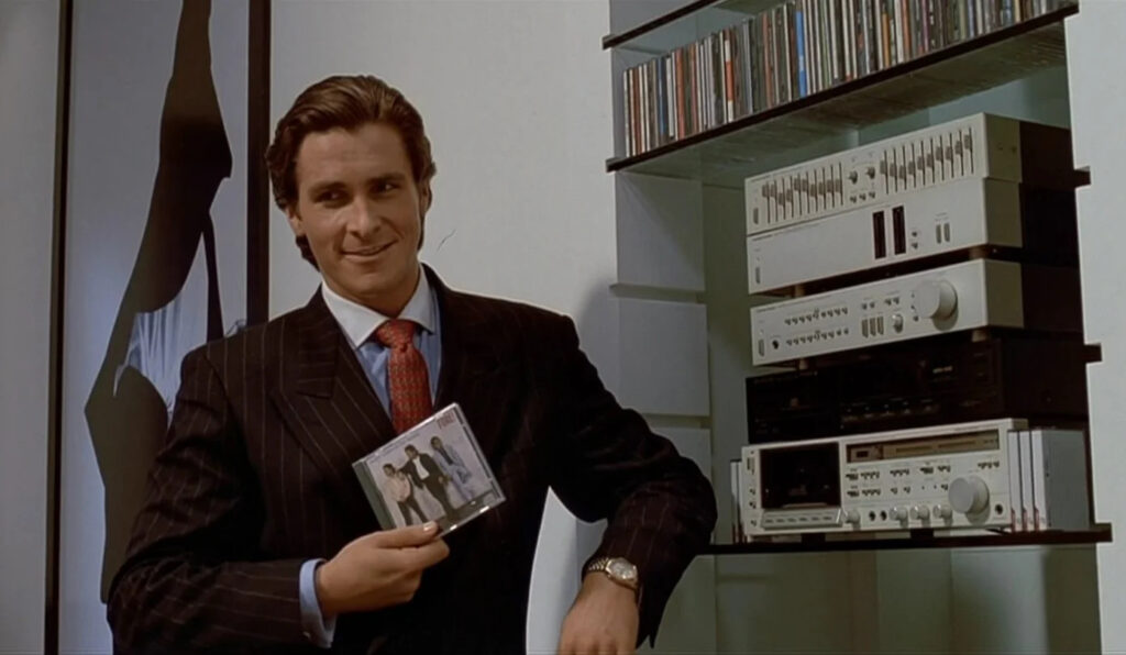 Patrick Bateman, a brunette white man wearing a pinstripe black suit with a blue button down and red patterned tie, holds up a CD. He is standing to the left of a CD stereo system.