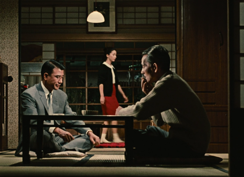 Two men sit at a table in the foreground. A woman in a black sweater and red skirt walks by in the background.