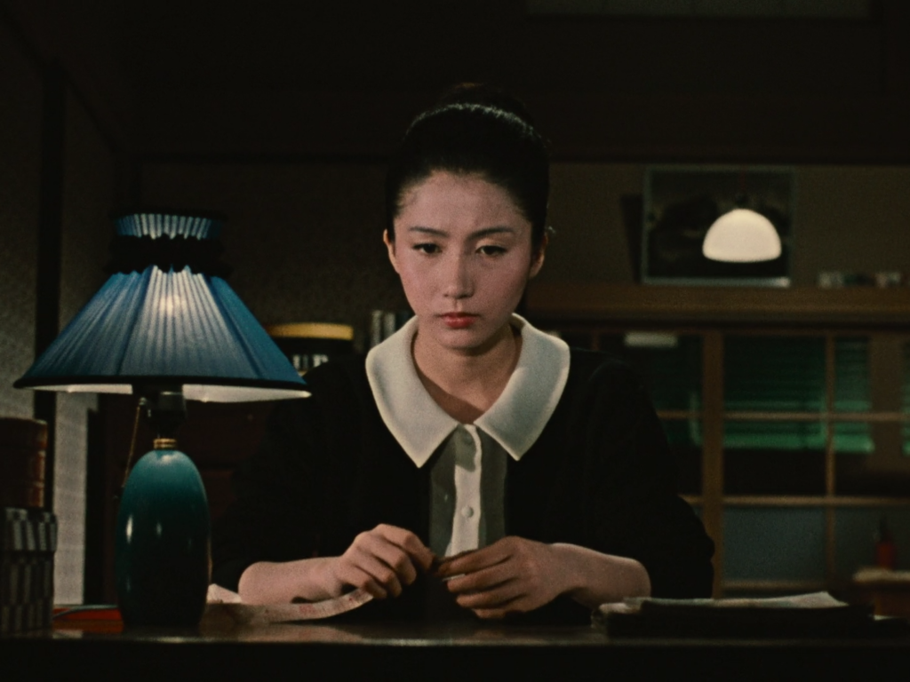 A woman in a black sweater sits at a desk appearing sad with a blue lamp in the foreground