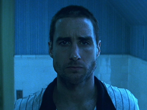 A man with a shaved head stands in a blue bathroom appearing sad.