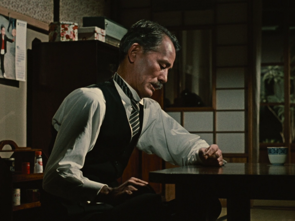 Man in a black vest and white shirt sits at a table and appears weary.