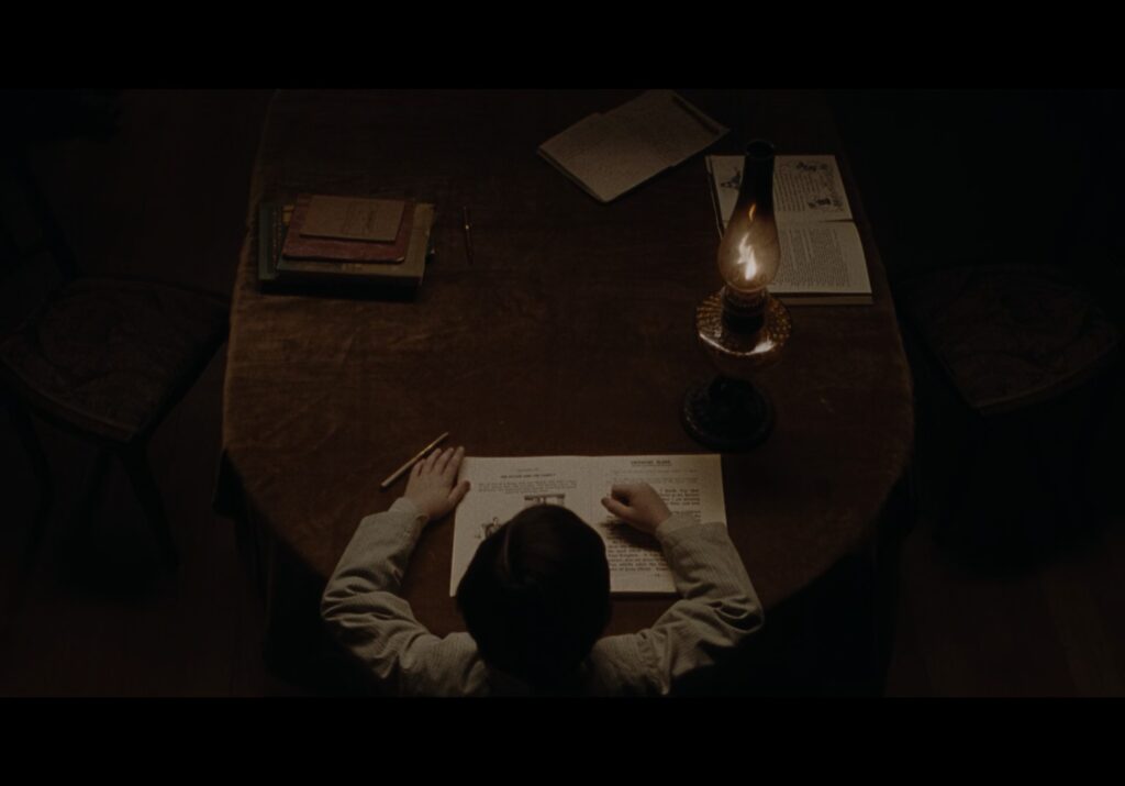 A young boy is studying a book at a wooden table by candlelight.