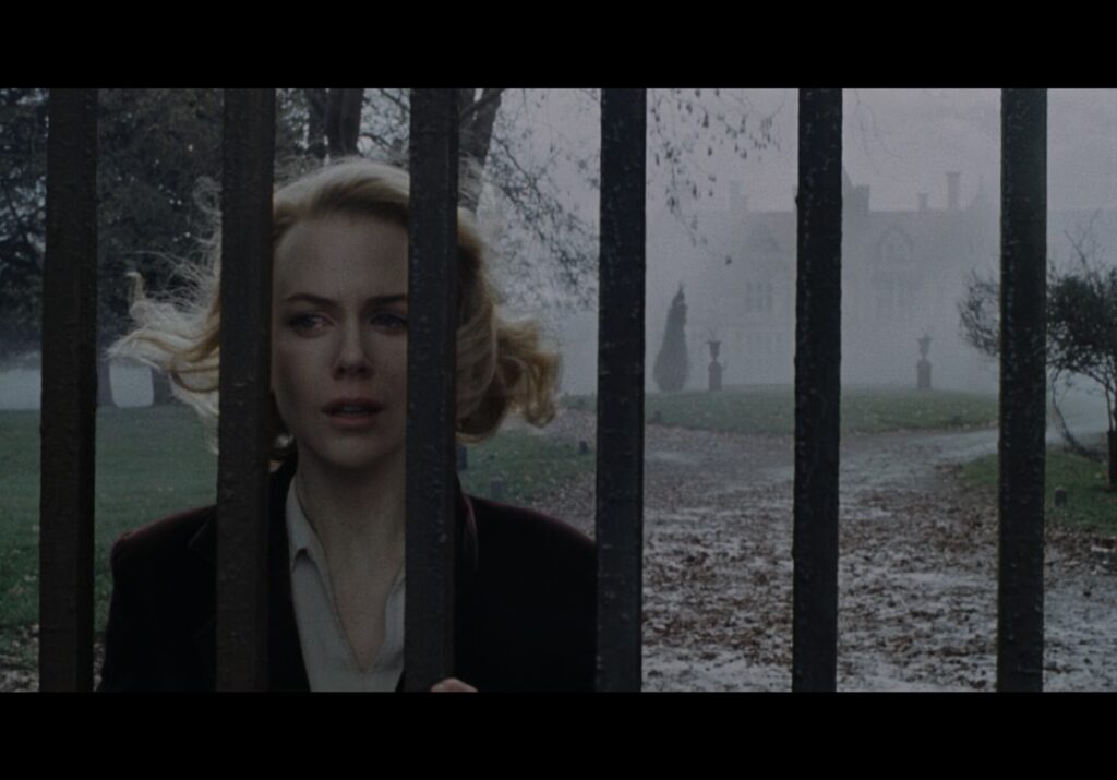  Nicole Kidman cries while looking through a metal gate in the fog front of a large mansion.