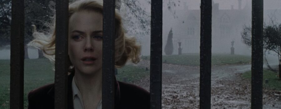 Nicole Kidman cries while looking through a metal gate in the fog front of a large mansion.