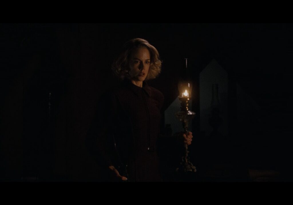 Nicole Kidman surrounded by darkness while holding an oil lamp.