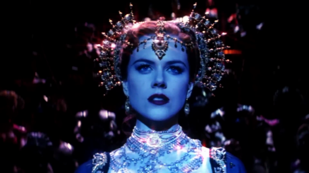 Nicole Kidman as Satine, a light-skinned ginger-haired woman illuminated by blue light, is wearing an elaborate head ornament and necklace and looks into the distance.