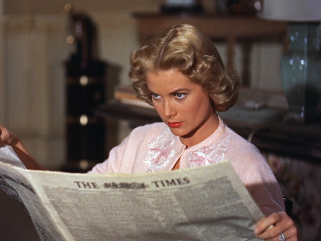 The wife, dressed in pink, looks up from her newspaper suspiciously