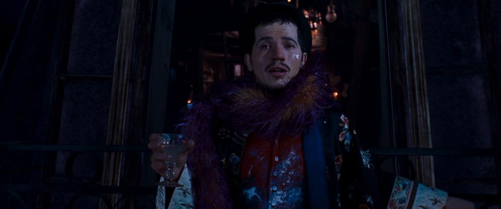 John Leguizamo as Toulouse, a light-skinned, dark-haired man with a small heart-shaped sticker on his face, sits on a building roof wearing a bright-colored feathered scarf and kimono, holding a glass of absinthe in his right hand.