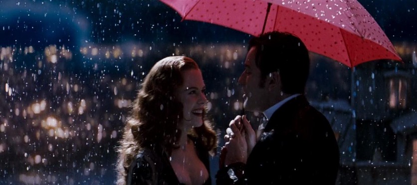 A man and woman smile at each other under a red umbrella in a rainstorm.