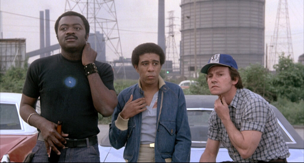 Actors Yaphet Kotto, Richard Pryor, and Harvey Keitel stand against a car drinking beers, looking concerned